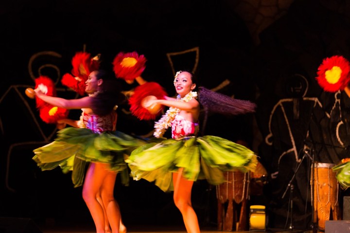 Hula dancers in green skirts and red feathers perform on stage.