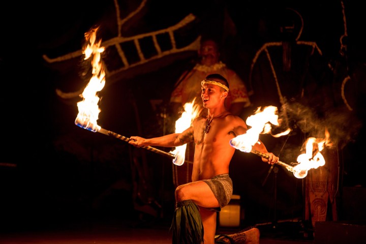 A performer kneeling while spinning two fire sticks at a nighttime show.