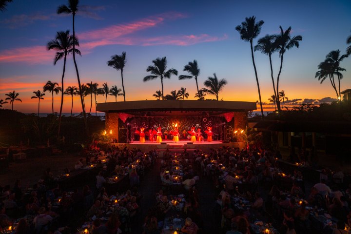 Outdoor evening luau with dancers on stage, palm trees, and vibrant sunset.
